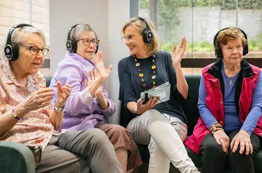 Elderly women and a middle-aged woman, all in headphones, enjoying a shared activity indoors.